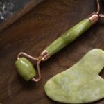 Gua Sha - From above of Jade roller and Gua Sha massage tool placed on wooden plate on black marble table with fresh twigs of rosemary