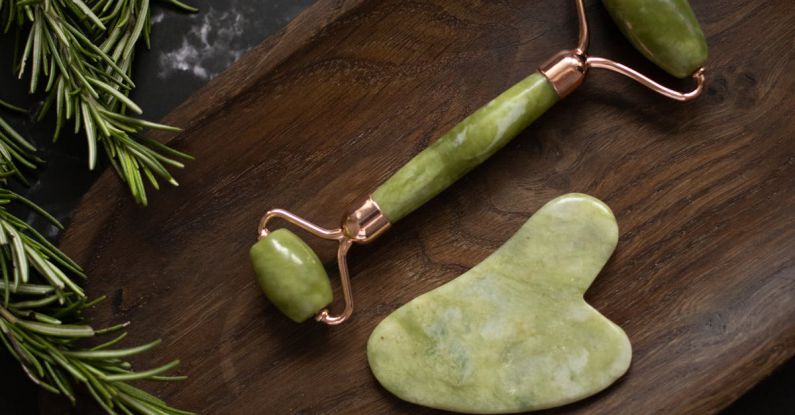 Gua Sha - From above of Jade roller and Gua Sha massage tool placed on wooden plate on black marble table with fresh twigs of rosemary